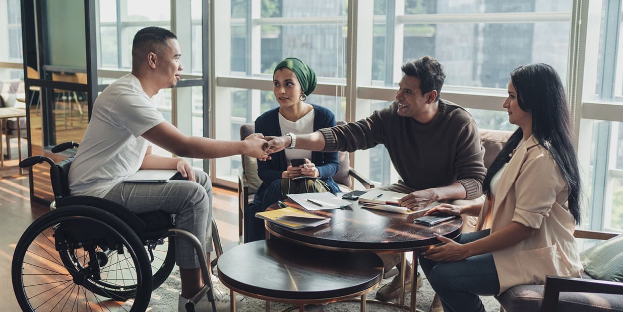 Een groep mensen zit zich rond een tafel en gaat met elkaar in gesprek. Er liggen blocnotes en pennen op tafel. Twee mannen schudden elkaar de hand over de tafel heen, één van hen zit in een rolstoel.